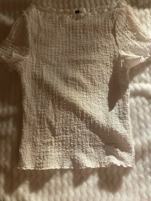 Vit kortärmad blus med struktur - Säljer en vit blus med korta ärmar och snyggt strukturerat tyg. Toppen har en rak passform och är tillverkad i ett lätt och luftigt material som passar perfekt till varmare dagar. Enkel och stilren design som är lätt att matcha.