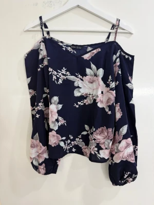 Blommig mörkblå blus  - Säljer en snygg blommig blus i mörkblått med rosa och gråa blommor. Blusen har v-ringad hals, offshoulder med smala axelband och långa puffärmar med resår vid ärmslut. Perfekt för dig som gillar romantiska detaljer och mjuka material.