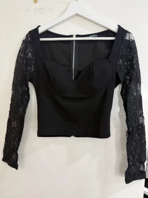 Svart korsettopp med spetsärmar - Säljer en svart korsettopp med långa spetsärmar och markerade kupor. Toppen har en snygg croppad passform och dragkedja baktill. Materialet är en mix av polyester och spets som ger en cool kontrast mellan kropp och ärmar.