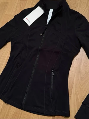 Svart Define Luon  jacka från Lululemon - Snygg svart träningsjacka från Lululemon med figurnära passform och hel dragkedja framtill. Jackan har två sidofickor med dragkedja, hög krage och diskreta sömmar som ger en sportig look. Materialet är mjukt och stretchigt! Skickas självklart i originalförpackning 😊💕