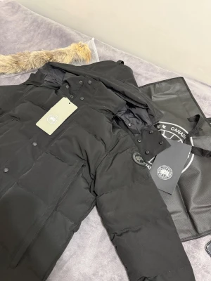 Canada Goose jacka  - Säljer en svart dunjacka från Canada Goose med huva och avtagbar fuskpäls. Jackan har flera knappar framtill, dragkedja och två stora fickor. Märkets patch sitter på ärmen och materialet är slitstark polyester. Perfekt för kalla vinterdagar.