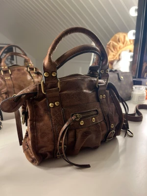 Brun handväska i skinn med detaljer - Säljer en brun hanböska i skinn med coola guld färgade nitar och dragkedja framtill. Väskan har både handtag och lång axelrem som går att ta av. Perfekt storlek för det viktigaste och ger en snygg vintage vibe till din outfit. Helt oanvänd!!💗💗