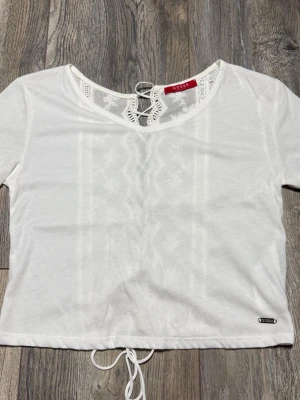 Vit spetsdetaljerad topp från Guess - Supersöt vit topp från Guess med korta ärmar och croppad passform. Toppen har spetsdetaljer och snörning baktill, samt ett litet metallmärke med Guess-logga vid fållen. Materialet känns mjukt och lätt, perfekt för varmare dagar.
