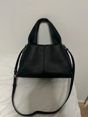 Svart axelväska i skinnimitation - Säljer en svart jätte fin handväska från Swedora. Det är den större modellen (storlek 30). Väskan är aldrig använd och är i nytt skick. Jätte bra med utrymme och bra material. Axelband ingår så det går att välja längd på väskan.
