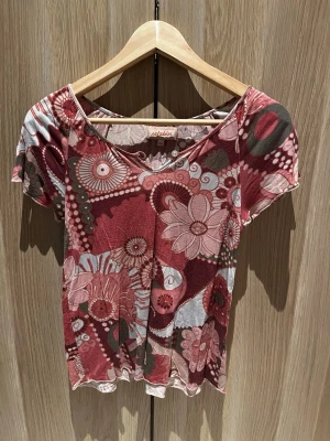 Blommig topp från Rabaldur - Unik topp från Rabaldur med blommigt mönster i vinröda, rosa och beige toner. Modellen har korta ärmar, rundad halsringning med knappdetalj och är gjord i ett mjukt, tunt material som känns skönt mot huden. Perfekt för dig som gillar färg och retrovibbar.