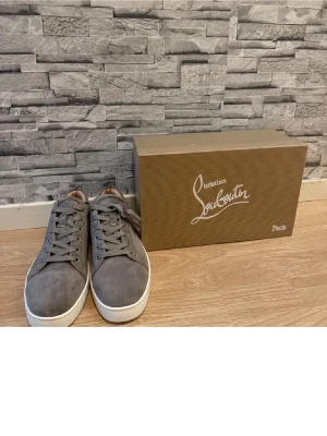 Grå sneakers från Christian Louboutin - Snygga grå sneakers från Christian Louboutin med klassisk röd sula och vit kant. Skorna är tillverkade i mocka och har rund tå, snörning och neutrala detaljer. Insidan är beige och de har en stilren, modern look som passar till många outfits. Kan sälja för ett billigare pris vid en snabb och smidig affär.