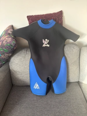 Blå och svart våtdräkt till barn  - Kortärmad våtdräkt i svart och blått från Target, perfekt för vattenaktiviteter. Dräkten har en cool surf-figur på bröstet och dragkedja i ryggen för enkel på- och avtagning. Tillverkad i tjockt syntetmaterial som håller värmen i vattnet. Köpt i Australien. Hals till gren 58 cm.