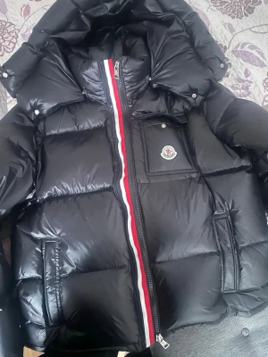 Svart Moncler pufferjacka med huva - Säljer en svart pufferjacka från Moncler med stor huva och ikonisk logga på bröstet. Jackan har en blank finish, dragkedja med röd och vit detalj, samt praktiska fickor. Perfekt för kalla dagar när du vill hålla stilen.