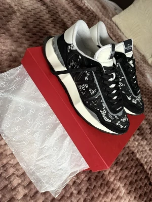 Valentino Garavani Rockrunner  - Säljer ett par svarta och vita Valentino Garavani Rockrunner sneakers med snygga blommiga spetsdetaljer. Skorna har snörning, rund tå och en chunky vit sula. Materialmix av mocka, spets och skinn ger en lyxig känsla. Kommer med originalkartong, extra skosnören och dustbag.