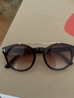 Solglasögon Tom Ford brun/guld - Säljer ett par snygga solglasögon från Tom Ford med rundad form och mörkbruna bågar. Glasen är tonade i brunt och bågarna har en elegant gulddetalj upptill samt på sidorna. Tillverkade i Italien och riktigt stilrena till sommaren. Serienummer tyvärr suddats bort, utöver det är de i jättebra skick.