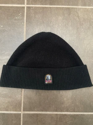Svart mössa från Parajumpers - Säljer en svart ribbstickad mössa från Parajumpers med klassisk patch framtill och röd etikett på insidan. Mössan är enkel och stilren, perfekt för kalla dagar och har en uppvikt kant. Passar dig som gillar snygga accessoarer med diskret logga.