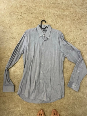 Ljusgrå slim fit skjorta från JOOP! - Säljer en ljusgrå skjorta från JOOP! i slim fit-modell. Skjortan har diskret fiskbensmönster, klassisk krage med knappar och vita knappar längs hela framsidan. Tillverkad i mjuk bomull och har långa ärmar med manschettknappar.