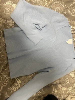 Ljusblå ribbad tröja från H&M - Säljer en ljusblå ribbad långärmad tröja från H&M. Tröjan har v-ringad hals och är gjord i mjuk bomullsmix som sitter skönt mot kroppen. Perfekt för dig som gillar stilrena och enkla plagg med lite struktur.