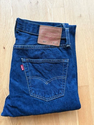 Levi's 501 mörkblå jeans W30 L30 - Klassiska Levi's 501 jeans i mörkblå tvätt med raka ben och femficksmodell. Jeansen har den ikoniska läderpatchen bak i midjan och röd Levi's-tag på bakfickan. Tillverkade i slitstarkt bomullsjeansmaterial och har knappgylf.