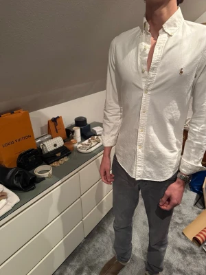 Vit slim fit skjorta från Ralph Lauren (Oxford) - Klassisk vit skjorta från Ralph Lauren i slim fit-modell. Skjortan har långa ärmar, knappar framtill och den ikoniska broderade loggan på bröstet. Tillverkad i mjuk bomull och passar perfekt till dig som vill se lite preppy ut.