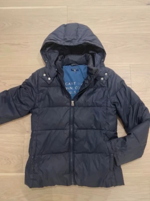 Mörkblå pufferjacka från Gant - Snygg mörkblå pufferjacka från Gant med huva och dragkedja framtill. Perfekt till vintern ny pris 1200kr för 13år 158 cm priset går att förhandla vid snabb affär