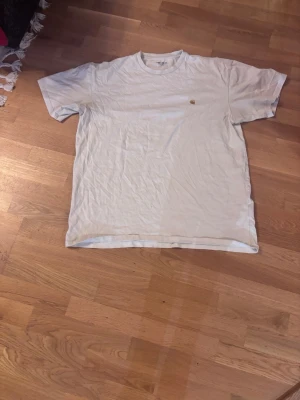 Vit t-shirt från Carhartt  - En vit vintage t-shirt från Carhartt med klassisk passform och rund hals. Liten gul Carhartt-logga på bröstet. Perfekt basplagg till garderoben, tillverkad i mjuk bomull för skön känsla hela dagen.