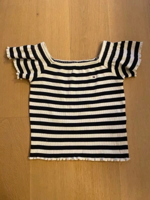 Randig topp från Tommy Hilfiger - Säljer en ribbad topp från Tommy Hilfiger med svart och vit randigt mönster. Toppen har korta, utställda ärmar och en fyrkantig ringning. Materialet är stretchigt och mjukt, perfekt för sommarens alla dagar. Liten logga broderad på bröstet.