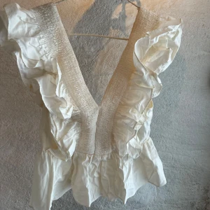 Vit volangtopp med djup v-ringning - Supersöt vit topp med djup v-ringning och breda volanger längs axlarna. Smockad upptill och med dragkedja i ryggen. Luftig och lätt, perfekt för varma dagar. Ger en romantisk och trendig vibe.