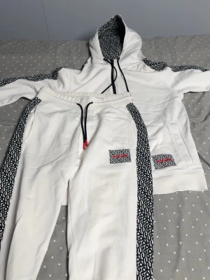 Vita mjukisbyxor med mönstrade detaljer - Säljer ett par vita mjukisbyxor från Gucci med svarta och vita grafiska mönster längs sidorna och på fickan. Byxorna har svarta snören med röda detaljer i midjan och är riktigt sköna för chill dagar. Matchar med hoodie för en clean och trendig look.