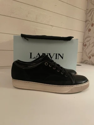 Svarta sneakers från Lanvin i mocka - Säljer ett par svarta Lanvin sneakers med ovandel i mocka och detaljer i blankt skinn. Skorna har vita, platta sulor och klassisk snörning. Tidlös och clean design som passar till det mesta. Kommer med dustbag. Storlek uk 8 men motsvar 42 men passar som 43. Lite tråkiga i hålen som syns på bilden 