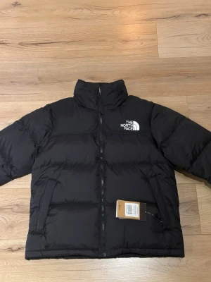 Svart pufferjacka från The North Face - Säljer en svart pufferjacka från The North Face, modell 1996 Nuptse. Jackan har hög krage, två sidofickor med dragkedja och tydlig logga på bröstet. Perfekt för kalla dagar och riktigt snygg till streetwear-looken.