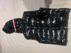 Svart dunväst från Moncler med huva - Säljer en glansig svart dunväst från Moncler med avtagbar huva. Västen har två dragkedjeförsedda fickor fram och vita/röda detaljer på huvan. Klassisk Moncler-logga på bröstet och ikonisk serietidningslapp på insidan. Perfekt för lager-på-lager under kyliga dagar.