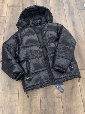  Polo Ralph Lauren jacka - Säljer en svart pufferjacka från Polo Ralph Lauren med huva och dragkedja. Jackan har två sidofickor med dragkedja, justerbar nederkant och den klassiska röda loggan på bröstet. Materialet är glansigt och fyllningen gör den perfekt för kalla dagar. Kvitto finns !