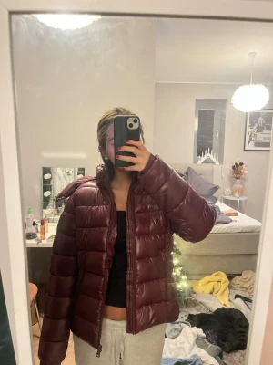 Mörkröd pufferjacka med dragkedja - Säljer en snygg mörkröd pufferjacka med hög krage och dragkedja framtill. Jackan är vadderad och har en glansig finish som ger en cool look. Perfekt för kalla dagar när du vill hålla dig varm och samtidigt se trendig ut.