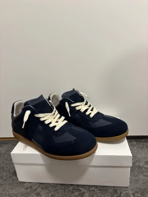 Maison Margiela sneakers marinblå 43 - Säljer ett par mörkblå sneakers med snygga kontraster i mocka och skinn. Skorna har vita snören och en klassisk gummisula. Perfekta för dig som gillar stilrena och tidlösa sneakers med en twist.