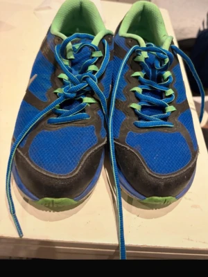 Nike blå sneakers med gröna detaljer - Säljer ett par blå Nike sneakers med gröna och svarta detaljer. Skorna har blå snörning, grönt foder och en sula i grönt, blått och svart. Ovandelen är i mesh och syntet, vilket gör dem lätta och ventilerande. Perfekta för träning eller vardagsbruk.