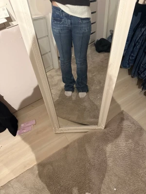 Blå bootcut jeans med fickor fram - Säljer ett par blå jeans med bootcut passform och stora fickor framtill. Jeansen har en klassisk blå tvätt och markerade sömmar längs benen. Materialet är jeans och de har en normal passform med midja som sitter bekvämt.