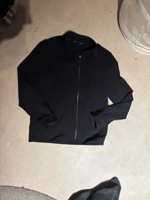 Svart overshirt från Zara - Säljer en svart overshirt från Zara med klassisk krage, två bröstfickor och hel dragkedja framtill. Jackan har långa ärmar med knapp vid mudden och är tillverkad i ett mjukt, lätt material som passar perfekt för lager på lager.