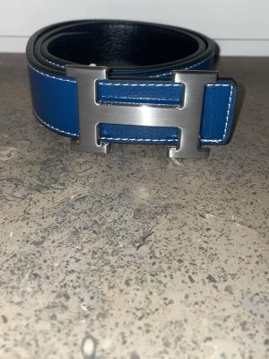 Blått bälte med metallspänne - Snyggt blått bälte med stort H-format metallspänne och vita kontrastsömmar. Bältet har en stilren look och är gjort i skinn. Perfekt accessoar för att lyfta din outfit.
