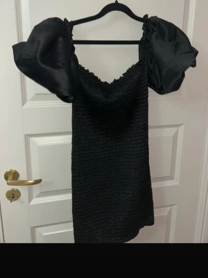 Svart kort klänning från Bik Bok - Supersnygg svart kort klänning från Bik Bok i storlek XS. Klänningen har puffiga korta ärmar i glansigt material och smockad kropp som ger en tight passform. Söt volangkant upptill och en elegant, stilren look. Perfekt för dig som vill sticka ut!