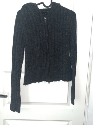 Mörkgrå ribbad hoodie med dragkedja - Mysig mörkgrå hoodie i ribbad stickad kvalitet med dragkedja framtill och huva. Tröjan har lång ärm och en något croppad passform med rå kant nedtill. Perfekt för lager på lager under kyliga dagar.