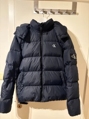 Mörkblå pufferjacka Calvin Klein Jeans - Snygg mörkblå pufferjacka från Calvin Klein. Finns ett litet hål på baksidan men syns inte alls och har bra helhetsintryck.