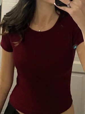 Vinröd kortärmad topp - Säljer en enkel och stilren vinröd topp med rund hals och korta ärmar. Modellen är figurnära och passar perfekt till jeans eller kjol. Materialet känns mjukt och stretchigt, vilket gör den bekväm att bära hela dagen.