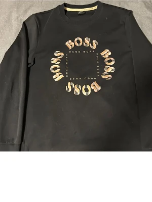 Svart sweatshirt från Hugo Boss - Säljer en svart sweatshirt från Hugo Boss med rund hals och tryck i beige och guld på bröstet där det står 'BOSS' i cirkel. Tröjan har långa ärmar och ribbade muddar vid hals, ärmslut och nederkant. Perfekt för en clean och stilren look.