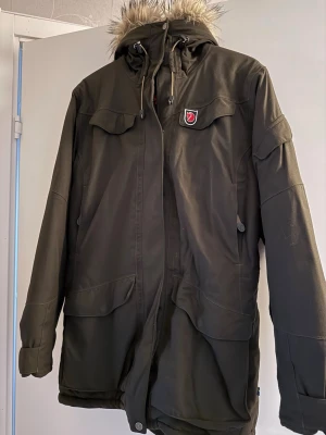 parkas från Fjällräven med fuskpäls- Brun S - Mörkbrun parkas från Fjällräven med huva och fuskpäls, stora fickor framtill och klassisk röd logga på bröstet. Jackan har dragkedja och knappar, samt justerbar huva. Perfekt för kalla dagar och outdoor.