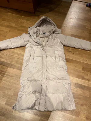 Beige lång pufferjacka från Zara - Lång beige pufferjacka från Zara med huva och dragkedja framtill. Jackan har två stora fickor på sidorna och quiltad design som ger en riktigt mysig vibe. Perfekt för kalla dagar när du vill hålla dig varm och ändå se snygg ut.