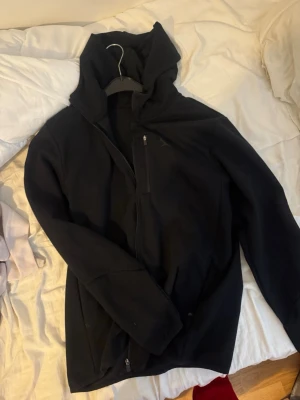 Svart vindjacka med huva - Säljer en svart vindjacka med huva och dragkedja framtill. Jackan har en enkel och stilren design med fickor och lång ärm. Perfekt för dig som vill ha en lätt och smidig jacka till vardags. Passar till många olika stilar.