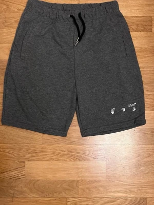 Mörkgrå shorts XL - Mörkgrå shorts i mjuk bomull med elastisk midja och svart snörning. Diskret logga och handmotiv broderat i vitt på ena benet. Perfekta för chill dagar eller streetwear-stil.