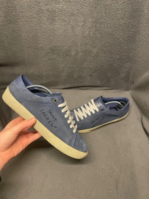 Blå Saint Laurent  - Säljer ett par snygga blå sneakers från Saint Laurent i mocka med vit sula och vita snören. Skorna har handskriven logga på sidan och klassisk låg profil. Perfekta för dig som vill ha en clean och stilren look med exklusiv känsla.