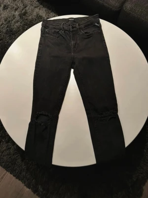 Svarta Replay jeans med slitningar - Säljer ett par svarta Replay jeans med slitningar över knäna. Modellen har klassisk femficksdesign och raka ben. Jeansen är tillverkade i ett mjukt bomullsmaterial med stretch för extra komfort. Perfekta för dig som gillar en edgy look.