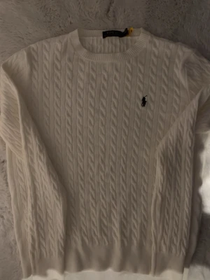 Vit kabelstickad tröja Polo Ralph Lauren - Kabelstickad vit tröja från Polo Ralph Lauren i storlek S. Klassisk rund halsringning och broderad marinblå logga på bröstet. Tröjan har lång ärm och ribbade muddar. Perfekt för dig som gillar tidlös och clean stil.