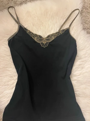 Svart linnetopp med guldbroderi Vero Moda - Säljer en svart linnetopp från Vero Moda i storlek S. Toppen har tunna axelband och snygga guldfärgade broderier längs ringningen och kanterna. Perfekt för dig som vill ha en enkel men ändå unik topp med fina detaljer.