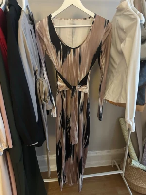 Långklänning med abstrakt mönster och knyt - Säljer en långklänning med coolt abstrakt mönster i beige, lila, svart och brunt. Klänningen har långa ärmar, rundad urringning och ett snyggt knyt i midjan som ger extra form. Materialet känns mjukt och stretchigt, perfekt för dig som gillar statement-plagg.