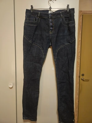 Mörkblå jeans från I Say med knappar - Snygga mörkblå jeans från I Say med fem dekorativa knappar i midjan och kontrastsömmar. Klassisk femficksmodell med raka ben och coola sömdetaljer framtill. Perfekta till vardagslooken och funkar året runt.