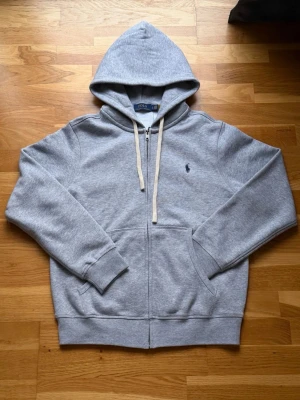 Ralph Lauren zip up grå S - Grå zip hoodie från Ralph Lauren i storlek S. Klassisk design med dragkedja, fickor fram och broderad logga på bröstet. Mjukt och bekvämt material, perfekt för vardagsbruk. I fint skick utan synliga skador eller fläckar.